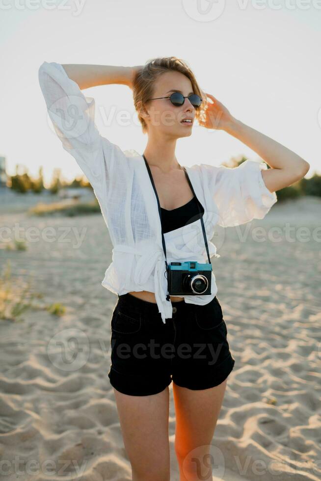 Elegant blond woman with retro camera posing on the beach near ocean. Beautiful sunlight. photo