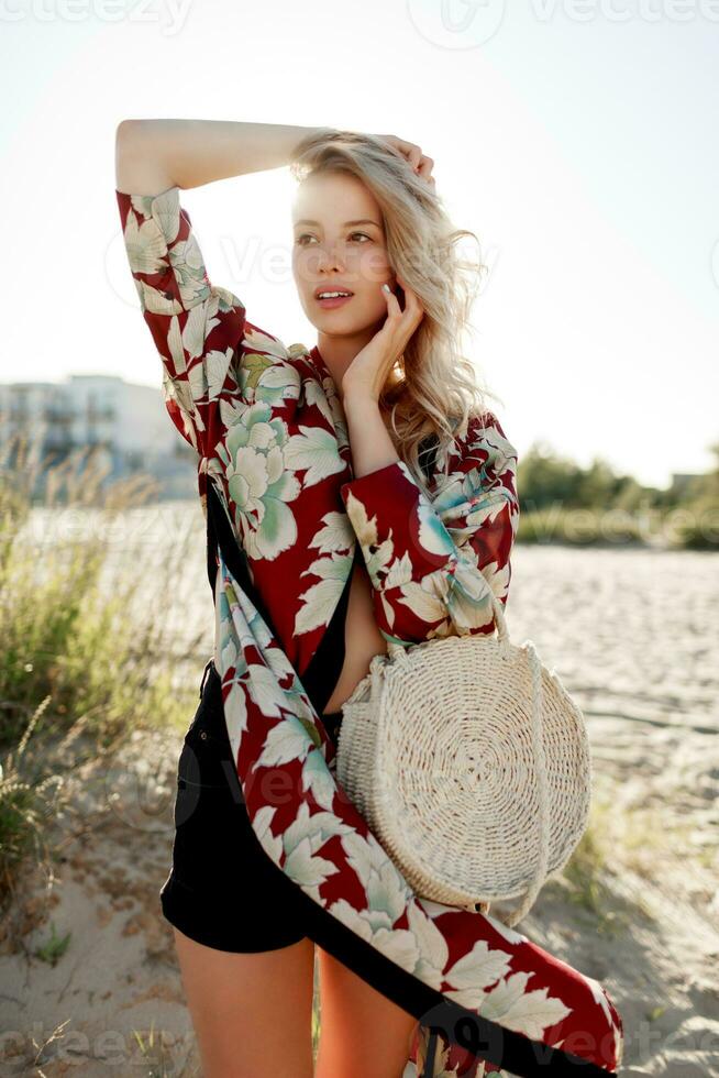 Gorgeous blond woman in trendy beachwear with floral print looking at camera on sunny beach. photo