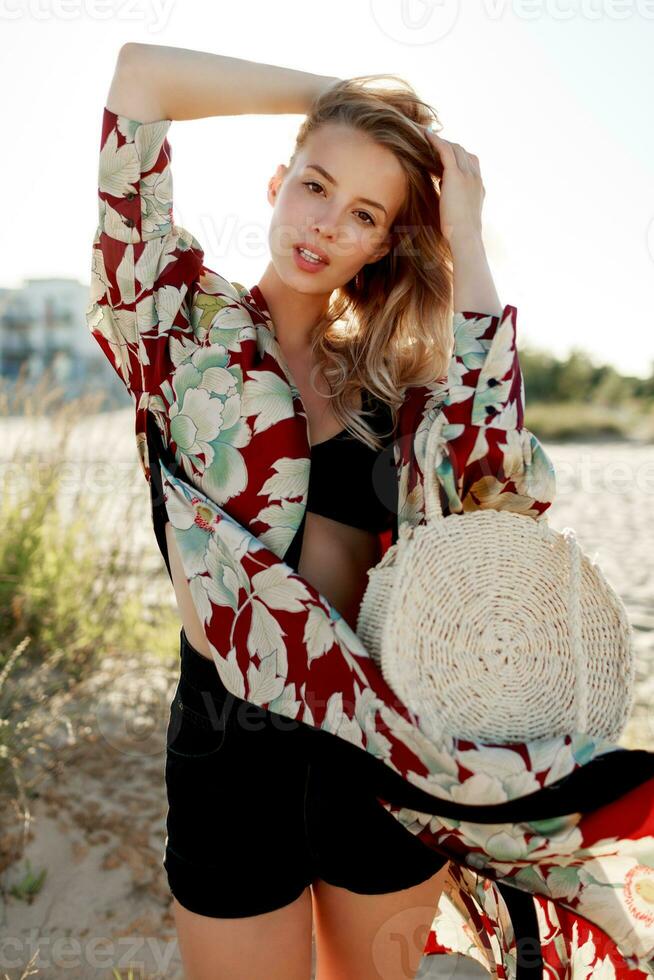 Gorgeous blond woman in trendy beachwear with floral print looking at camera on sunny beach. photo