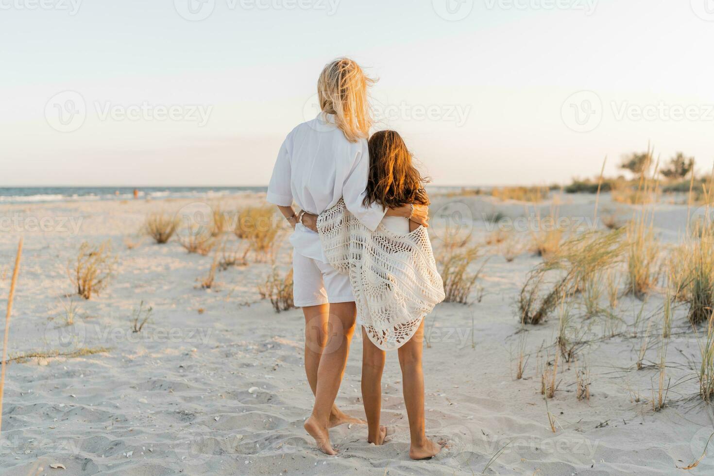European  mom with cute little daughter posing on sunny beach. Wearing light boho summer clothes. Sunset light. Family vacations concept. photo