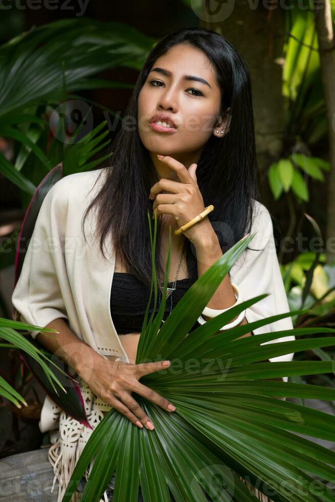 bonito asiático niña posando en tropical jardín, participación grande palma hoja. foto