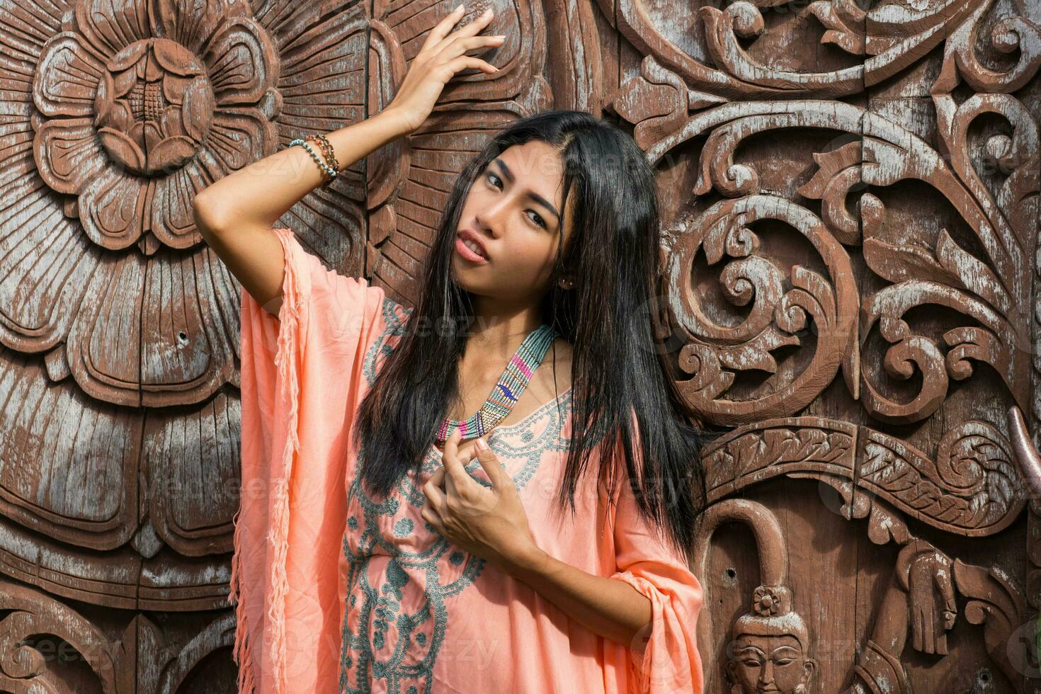 Stunning asian woman in boho ethnic dress posing over wood ornamental background. Looking at camera. photo