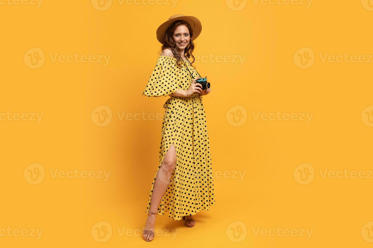 Full lenght photo of Travelling woman in straw hat and summer dress posing with retro camera over yellow backgrond in studio.