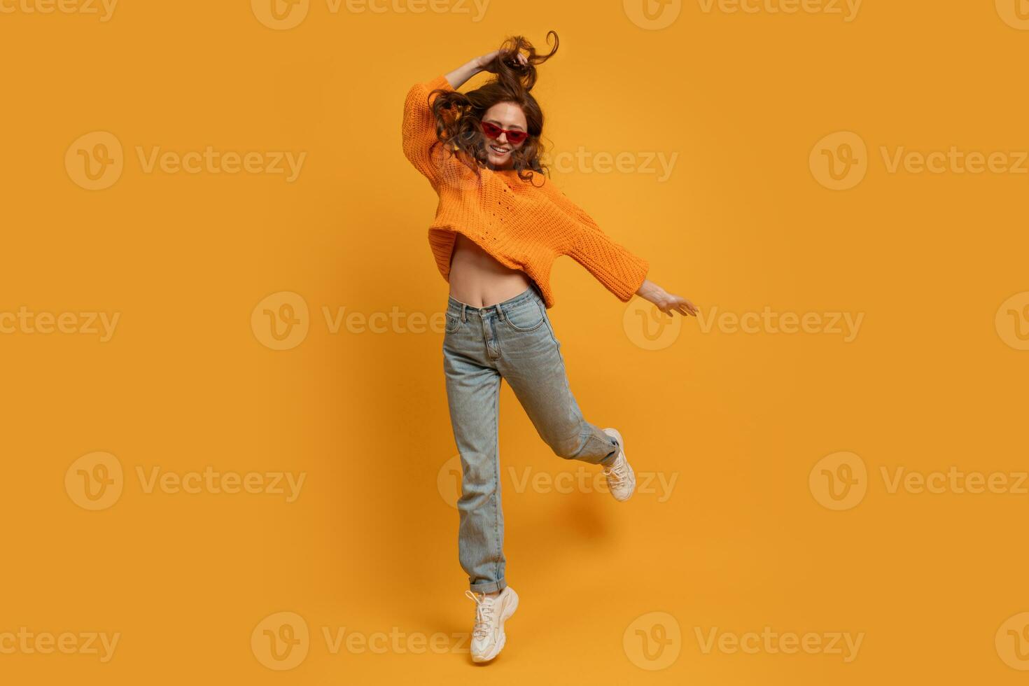 Laughing stylish red head  woman in orange sweater and blue jeans  jumping  over yellow  background.  Full lenght. photo