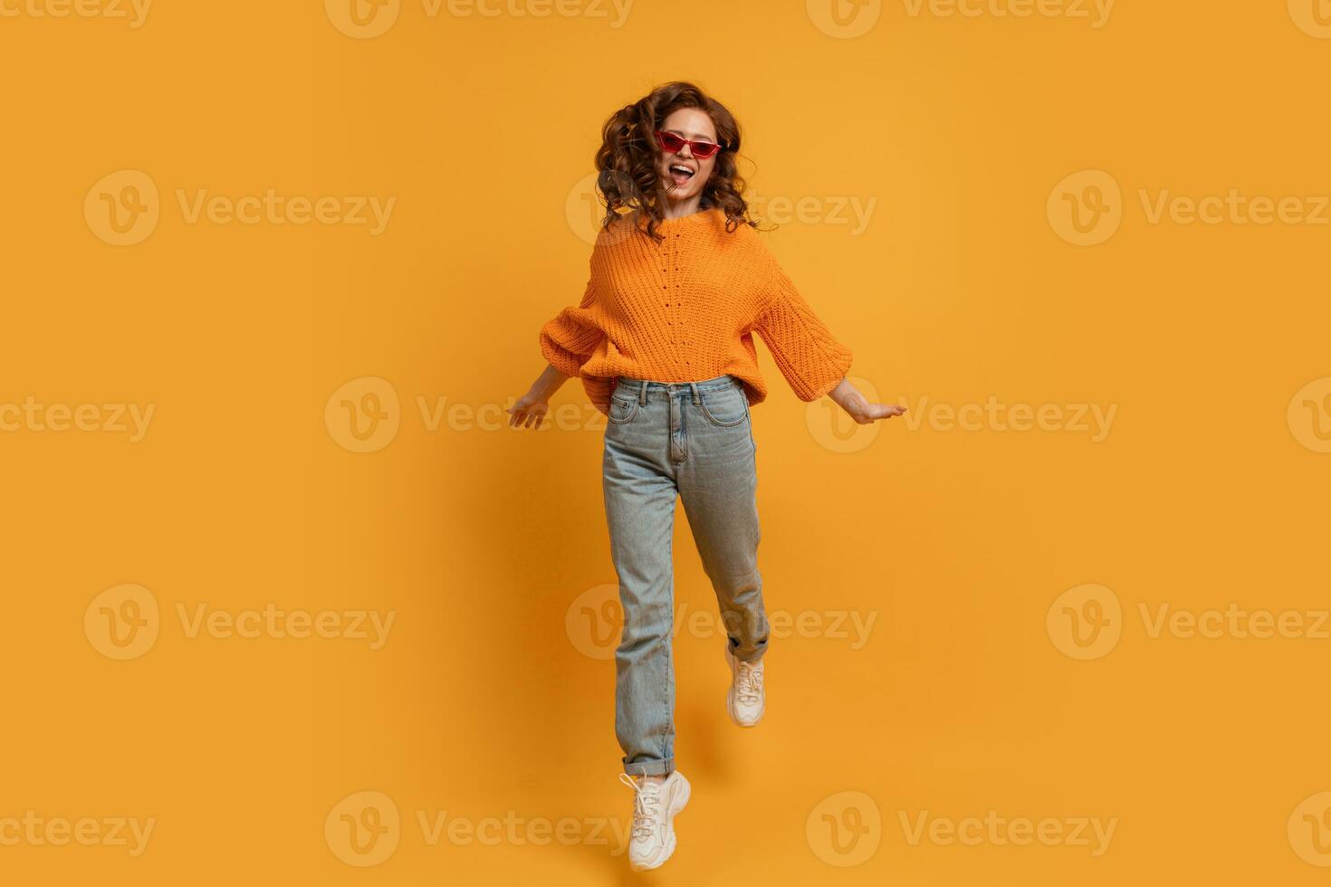 Laughing stylish red head  woman in orange sweater and blue jeans  jumping  over  yellow background.  Full lenght. photo