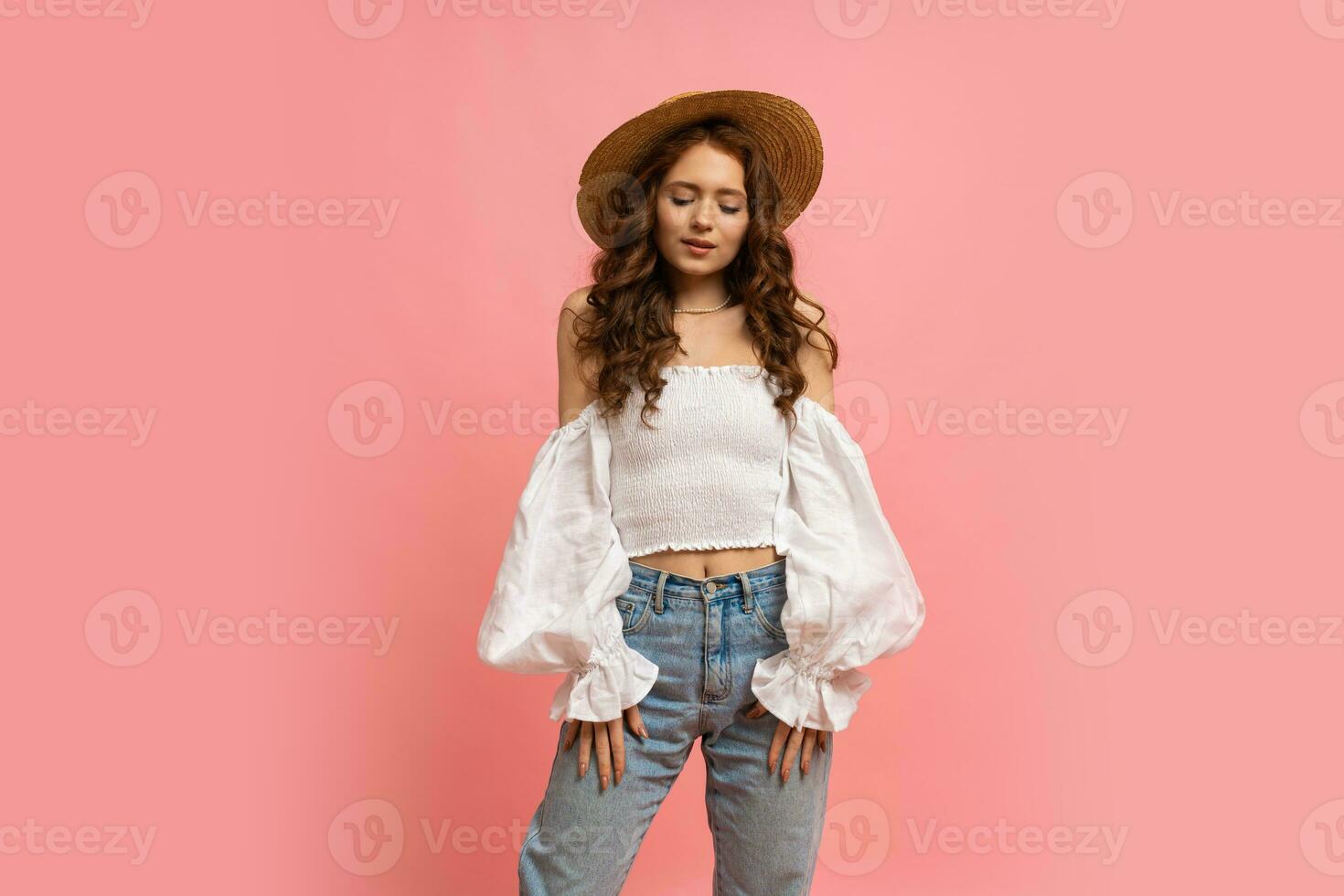Fashion studio photo of elegant red haired  girl in straw hat and summer outfit posing on pink background.