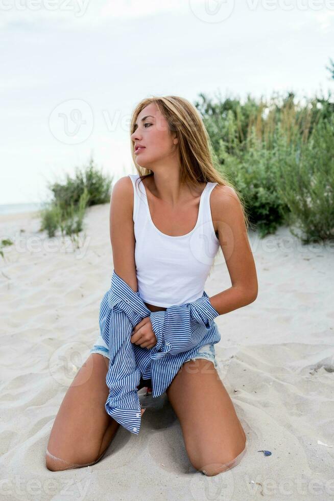cerca arriba retrato de rubio seductor niña con largo brillante ondulado pelos en verano playa disfrutando vacaciones. a rayas blusa, pantalones bermudas. Paja sombrero. foto