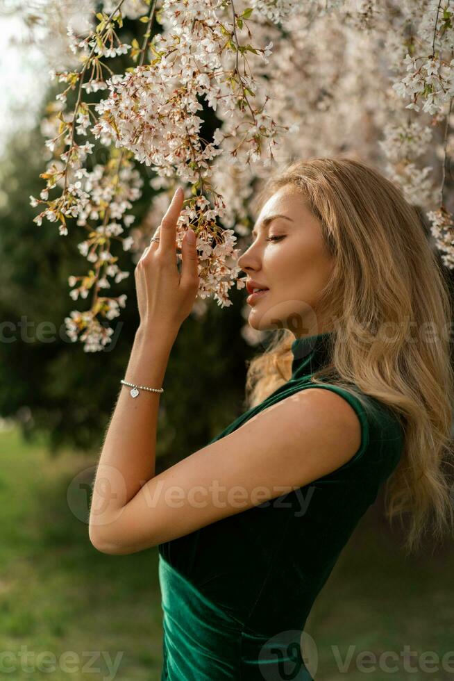 Fashion portrait of beautiful young lady with stylish makeup and wavy  long  hair. Big lips, soft skin and beautiful eyes. Wearing  velvet green  dress.  Posing near nlooming tree. photo