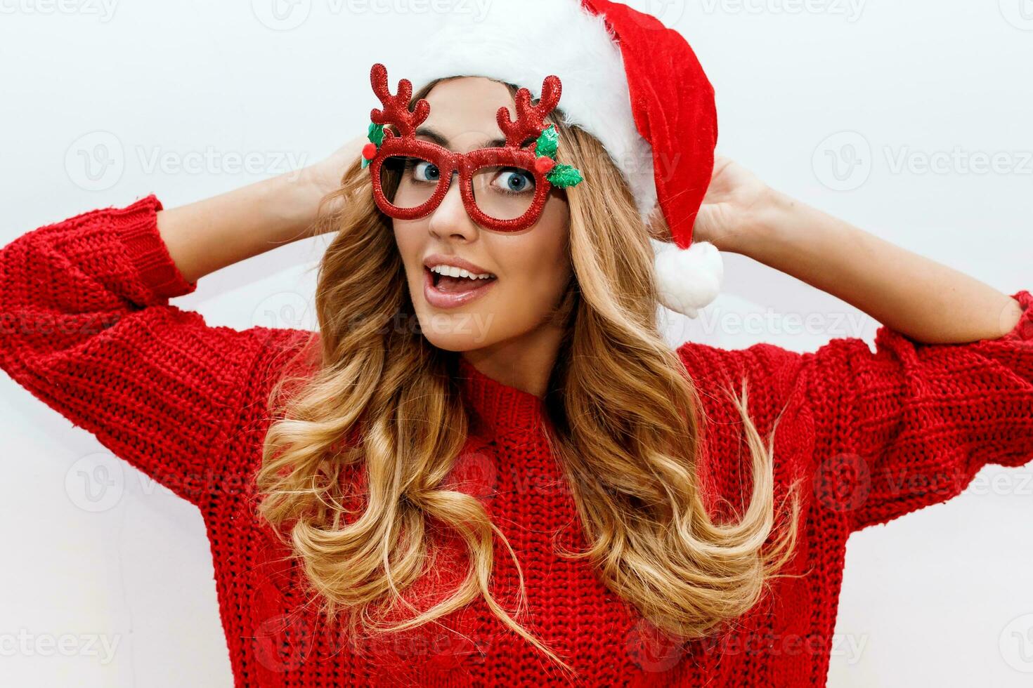 Joyful carefree blond woman in cute masquerade glasses and new year hat in red knitted sweater posing on white background . Isolate. Christmas and new test party concept. photo