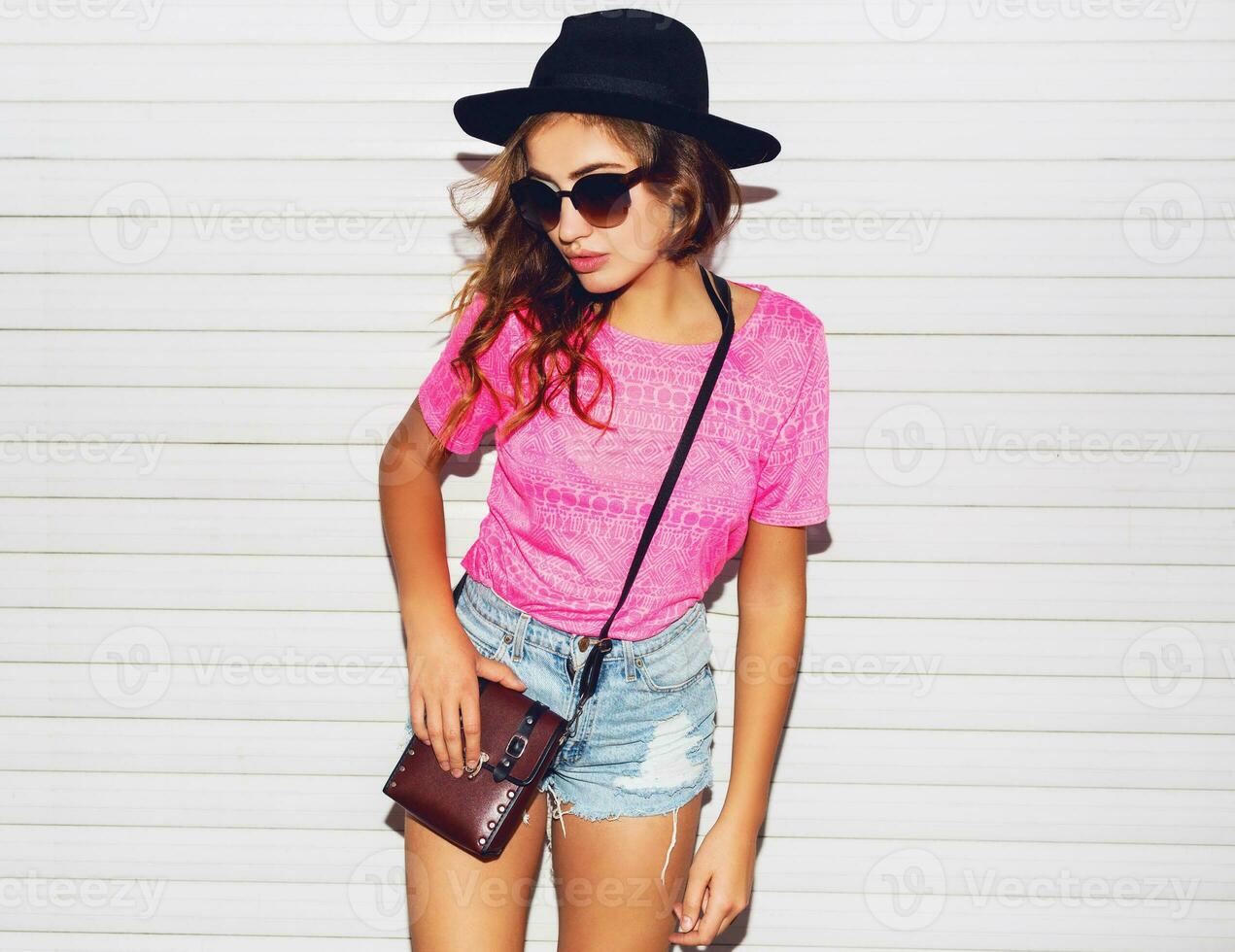 elegante noche destello Moda retrato de de moda casual joven mujer en rosado neón camiseta , negro sombrero, elegante camisas posando cerca blanco urbano pared a lo largo . foto