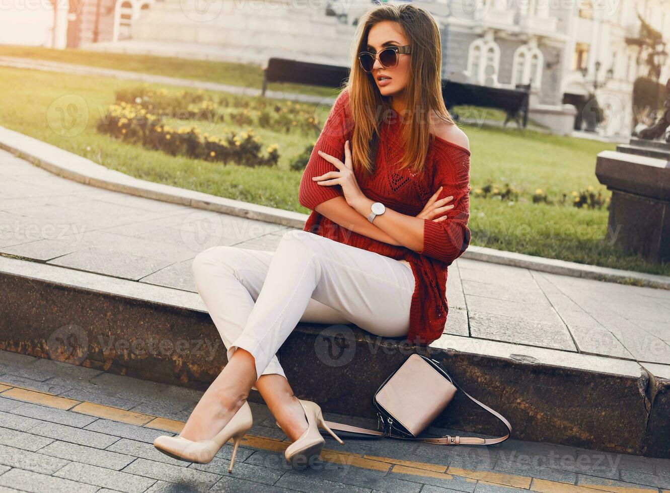 Pretty stylish  woman in trendy sunglasses and red sweater sitting and   flirting . Outdoor street style portrait. Cheerful woman wearing  heels and white jeans. photo