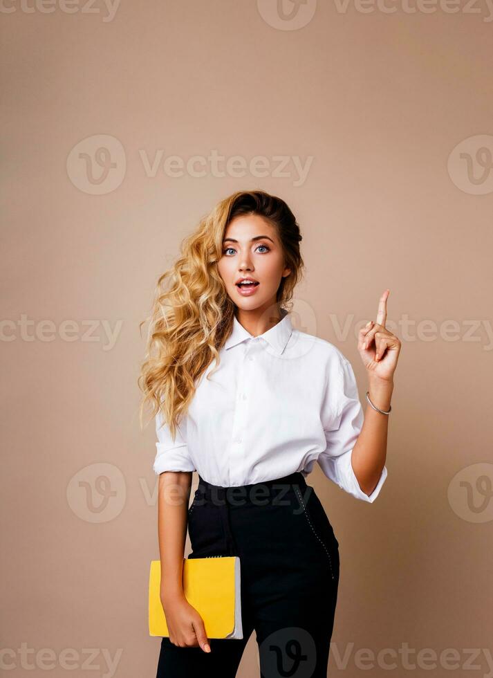 Blond business woman pointing up and looking at camera on beige background. Wearing stylish workwear. Copy space fo text.  Surprise face. photo