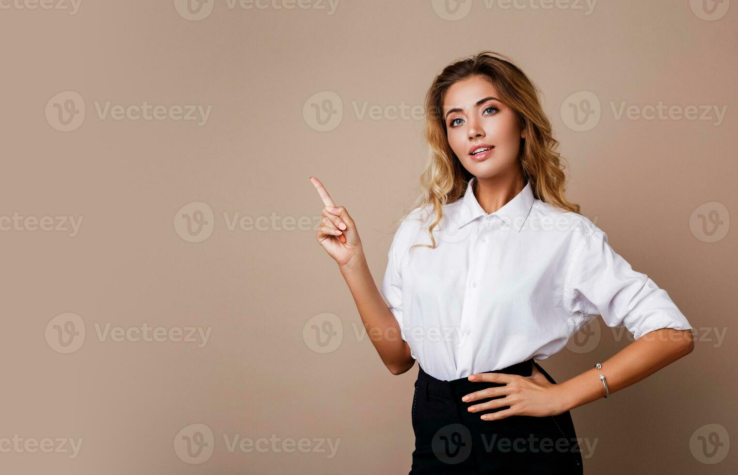 Blond business woman pointing up and looking at camera on beige background. Wearing stylish workwear. Copy space fo text. photo