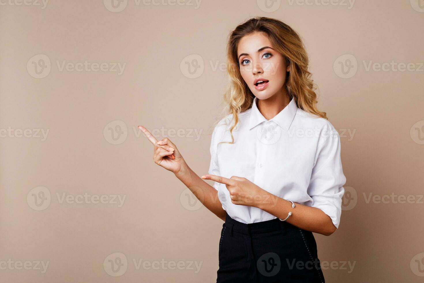 Blond business woman pointing up and looking at camera on beige background. Wearing stylish workwear. Copy space fo text. photo