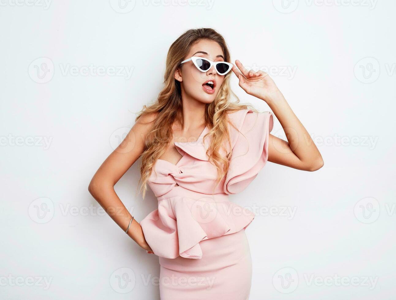 Beautiful blonde woman with surprised face standing over white background in studio. Wearing fashionable stylish dress. Emotional face. photo