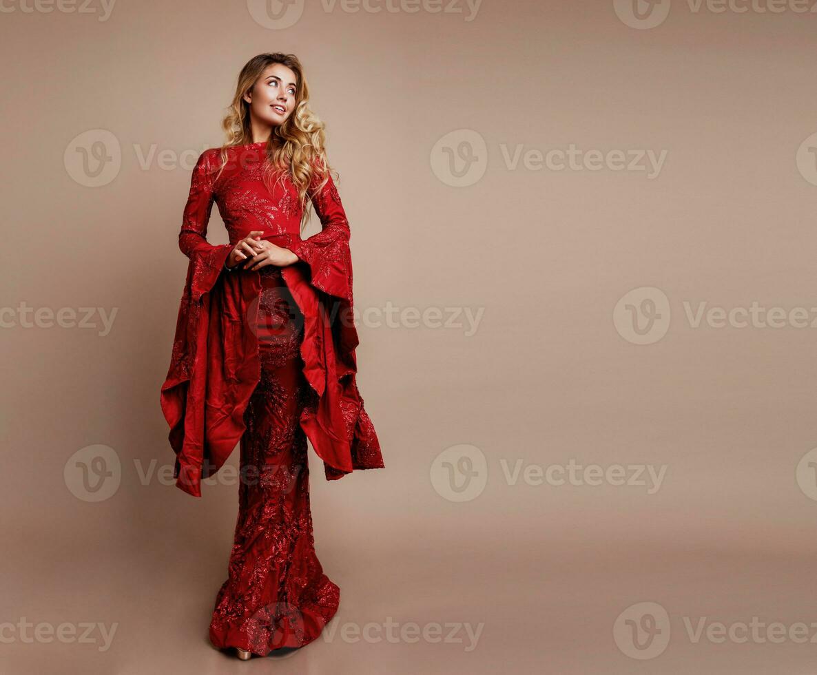 Graceful Blonde woman in elegant new year dress posing on studio.  Unusual wide sleeves. Wavy hairs. Full height. Beige background. Copy space for text. photo