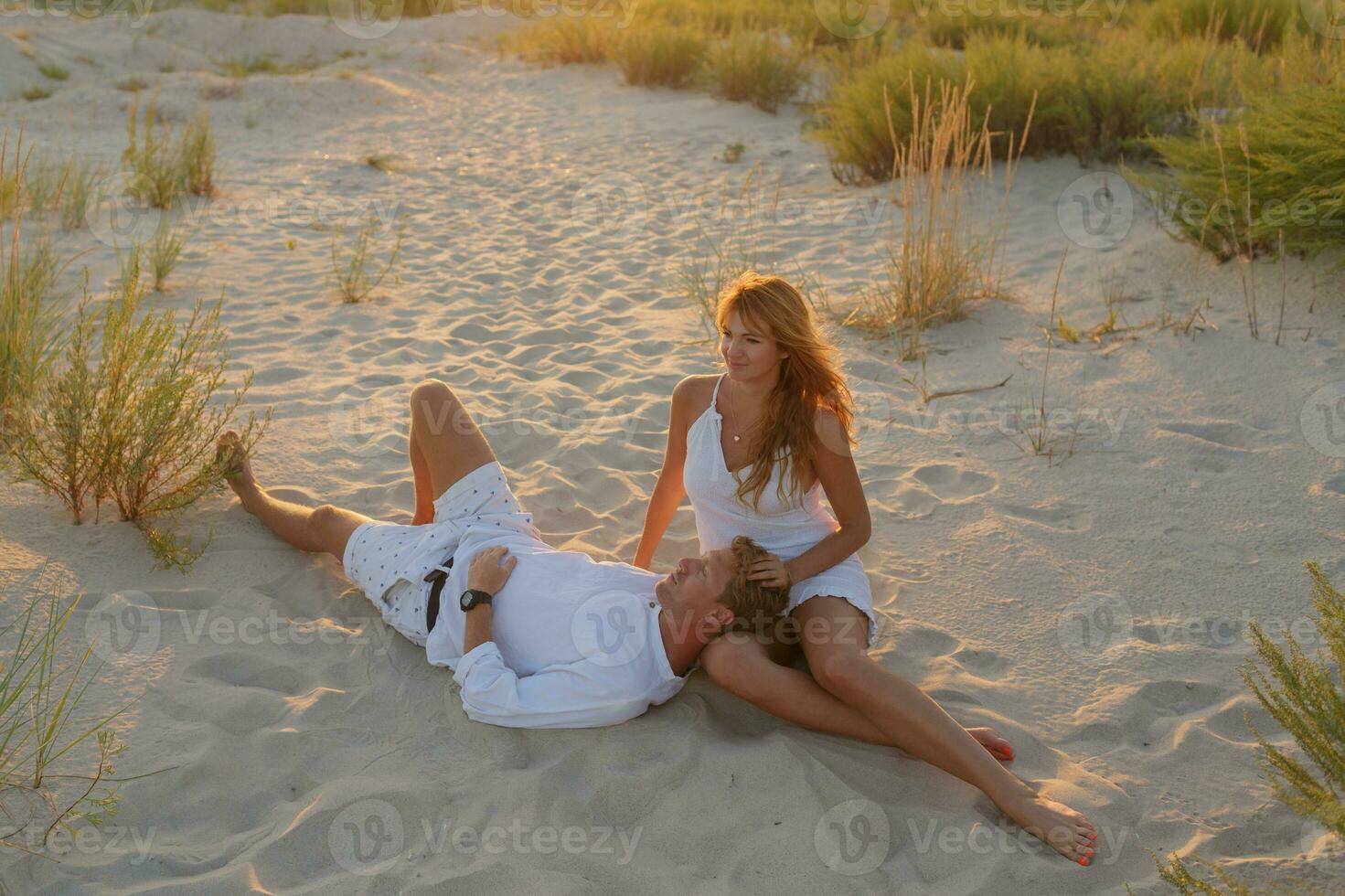 verano retrato de joven elegante Pareja sentado en el playa en soleado noche . foto