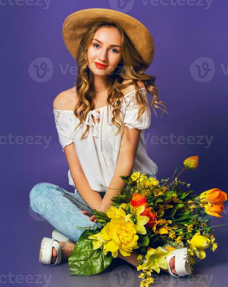portrait of  beautiful blonde young lady in stylish straw summer hat holding   colorful spring flower bouquet   near  purple wall background. photo