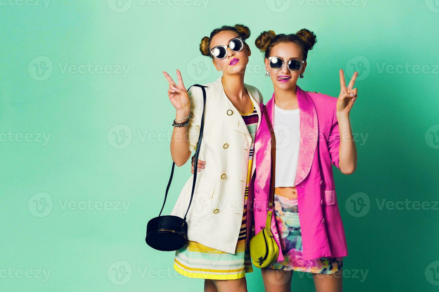 Fashion studio image of two young women in stylish casual  spring outfit   having fun, show  tongue. Bright trendy   colors, stylish hairstyle  with buns , cool sunglasses. Friends portrait. photo