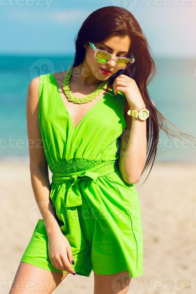 Fashion  outdoor summer portrait of pretty young  brunette beautiful woman in  cool sunglasses posing on the sunny tropical  beach. photo