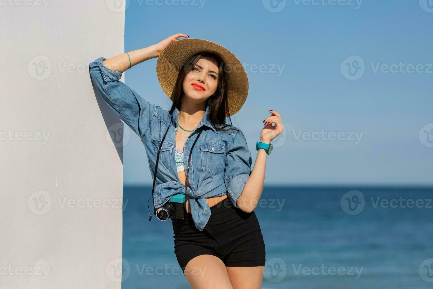 Happy  woman with perfect figure in sexy shorts and straw hat posing on the beach. Blue sky and sea on background. photo