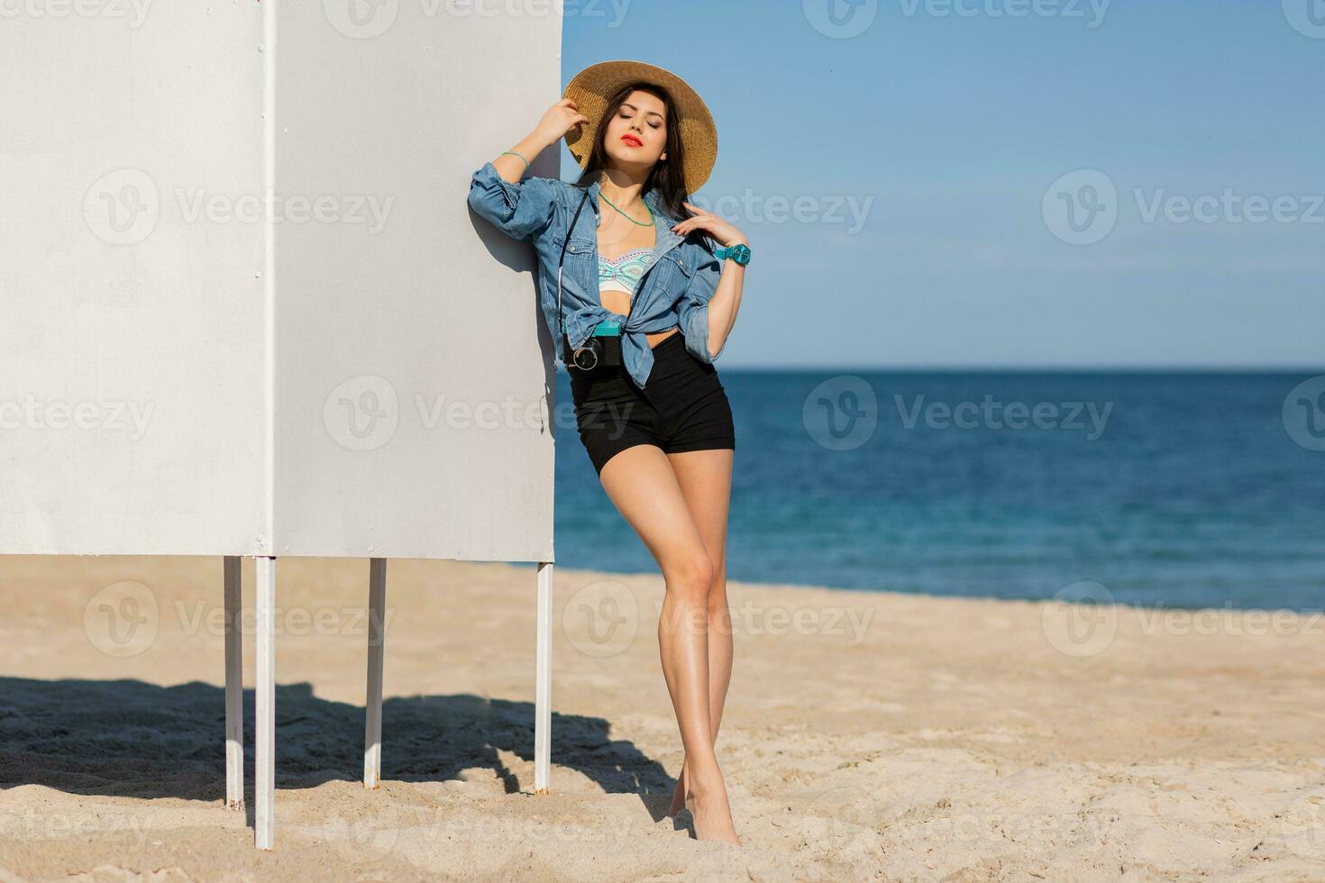 maravilloso mujer con Perfecto figura en sexy pantalones cortos y Paja sombrero posando en el playa. azul cielo y mar en antecedentes. lleno largo. foto