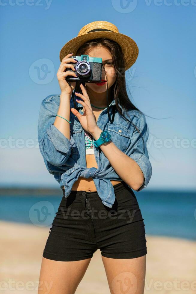 sexy mujer con Perfecto bronceado cuerpo, lleno rojo labios y l largo piernas posando en el tropical soleado playa. vistiendo cosecha parte superior , pantalones cortos y Paja sombrero. brillante soleado colores. foto