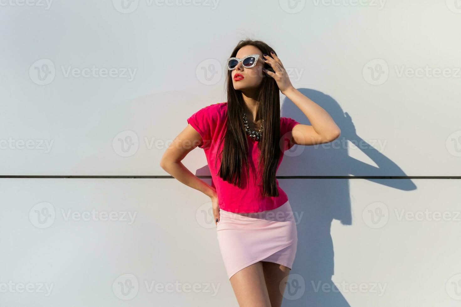 elegante mujer en verano ropa y Gafas de sol posando terminado blanco urbano antecedentes. foto