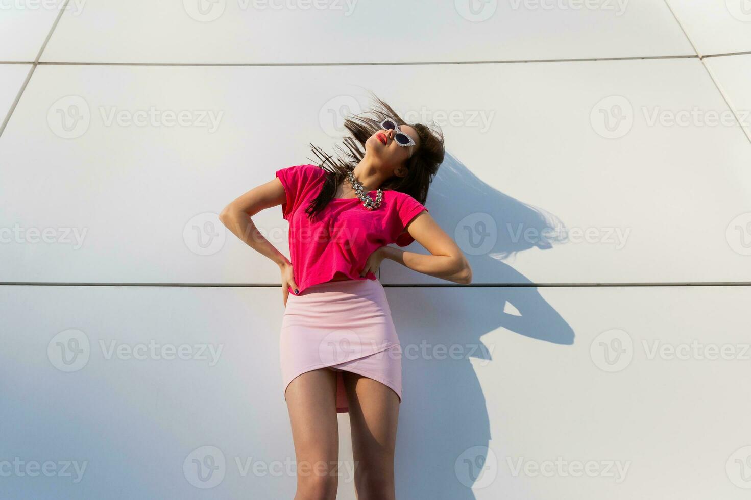 Fashionable brunette woman in summer clothes  and sunglasses posing over white urban background. Dancing and having fun. photo