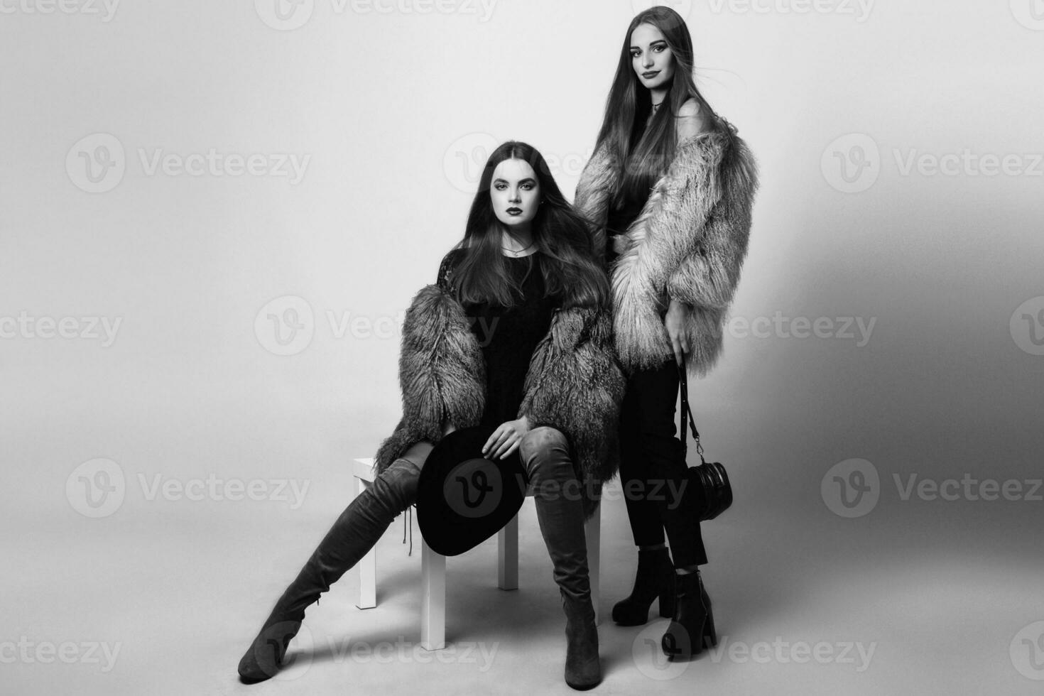 two  beautiful fashion models in  winter clothes  furs  posing on  gray background. Wearing stylish fluffy coat , black jeans, evening dress and hat. Black and white. photo