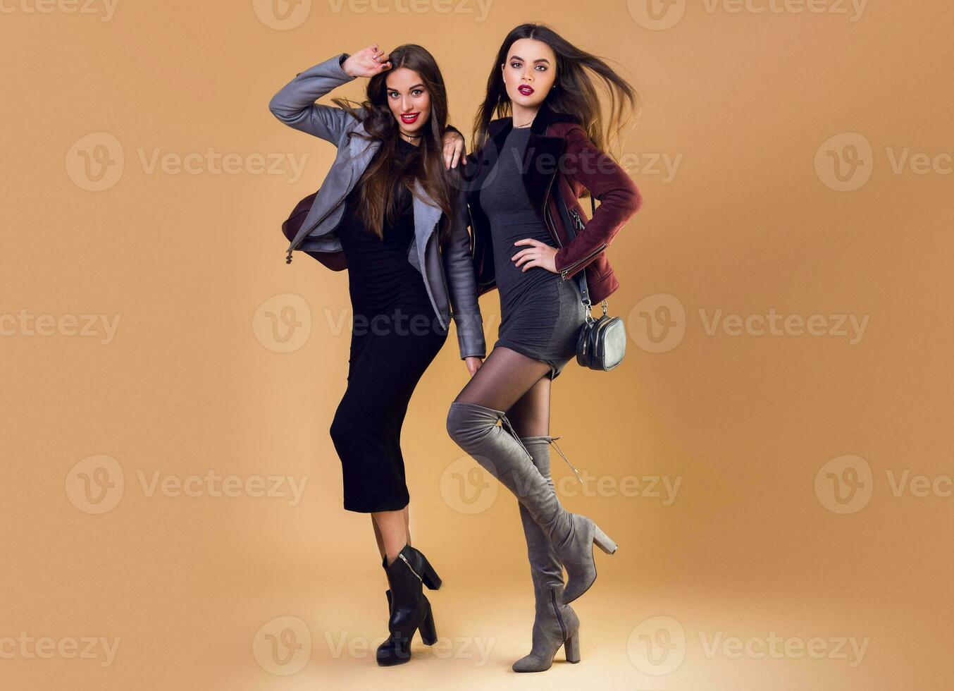 Glamorous  pretty two girls posing over beige   background ,  wearing casual winter jacket and dress. high heels.  Windy hairstyle. Full length image. photo