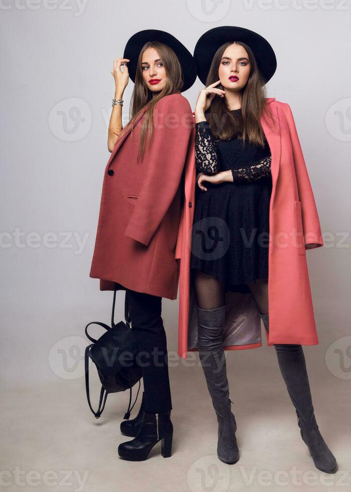 Full height   studio image of two fashionable sexy woman in casual trendy spring coat,  boots with heels , black hat, stylish hand bag. photo