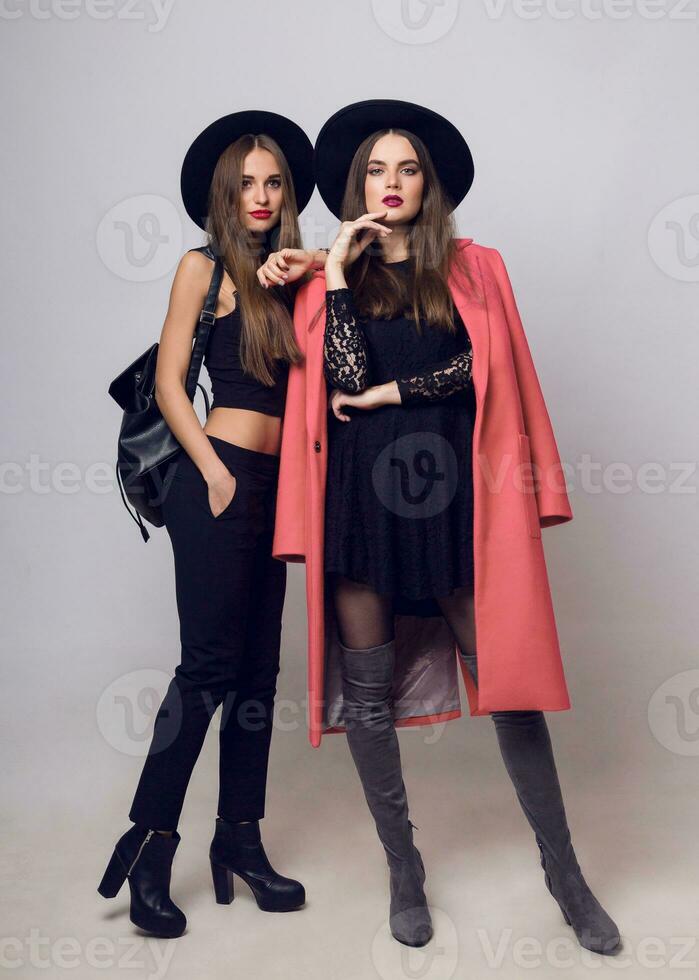 Full height   studio image of two fashionable sexy woman in casual trendy spring coat,  boots with heels , black hat, stylish hand bag. photo