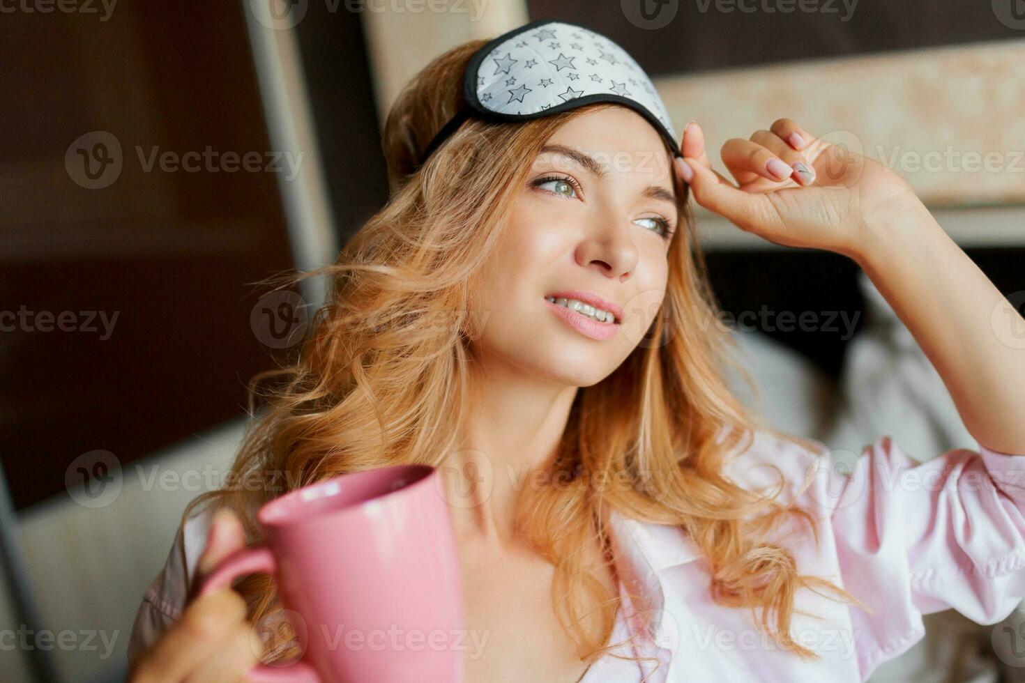 Pretty woman with candid smile posing in eye mask  at home and drinking hot tea. Cozy home atmosphere. photo