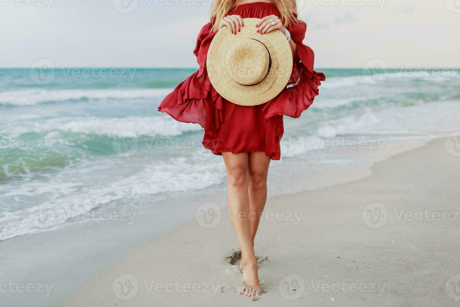 White  ginger  woman in stylish dress  holding straw hat and  posing  on the beach near ocean. Blu sky. Windy weather photo