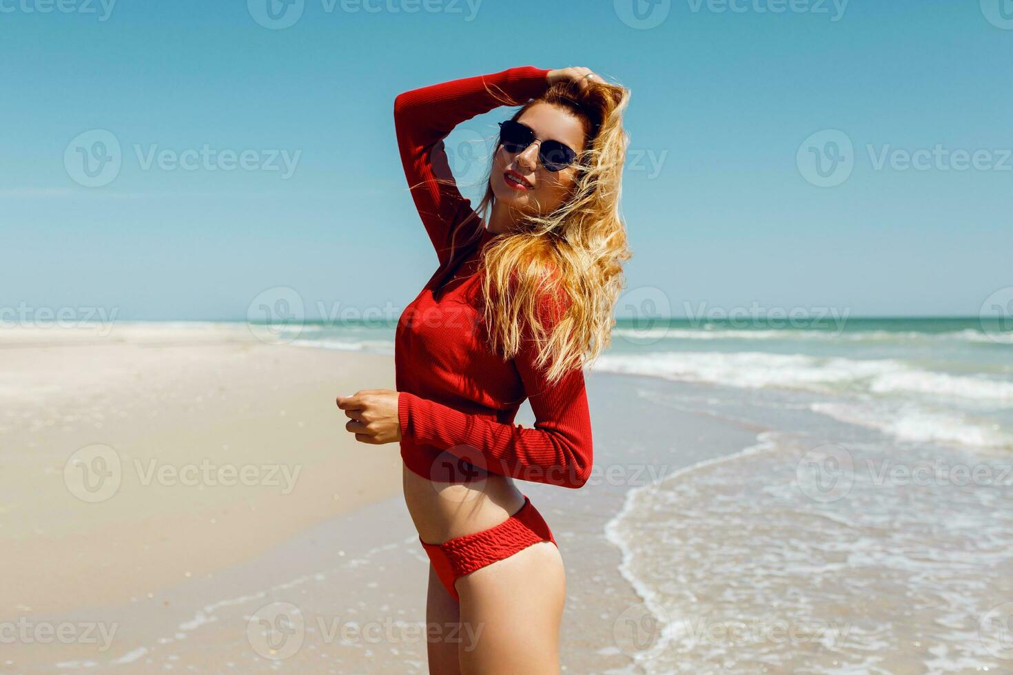 Close up summer portrait of  dreamy happy blond woman in red bikini posing on empty amazing beach near ocean. Exotic island.  Freedom and vacation concept. photo