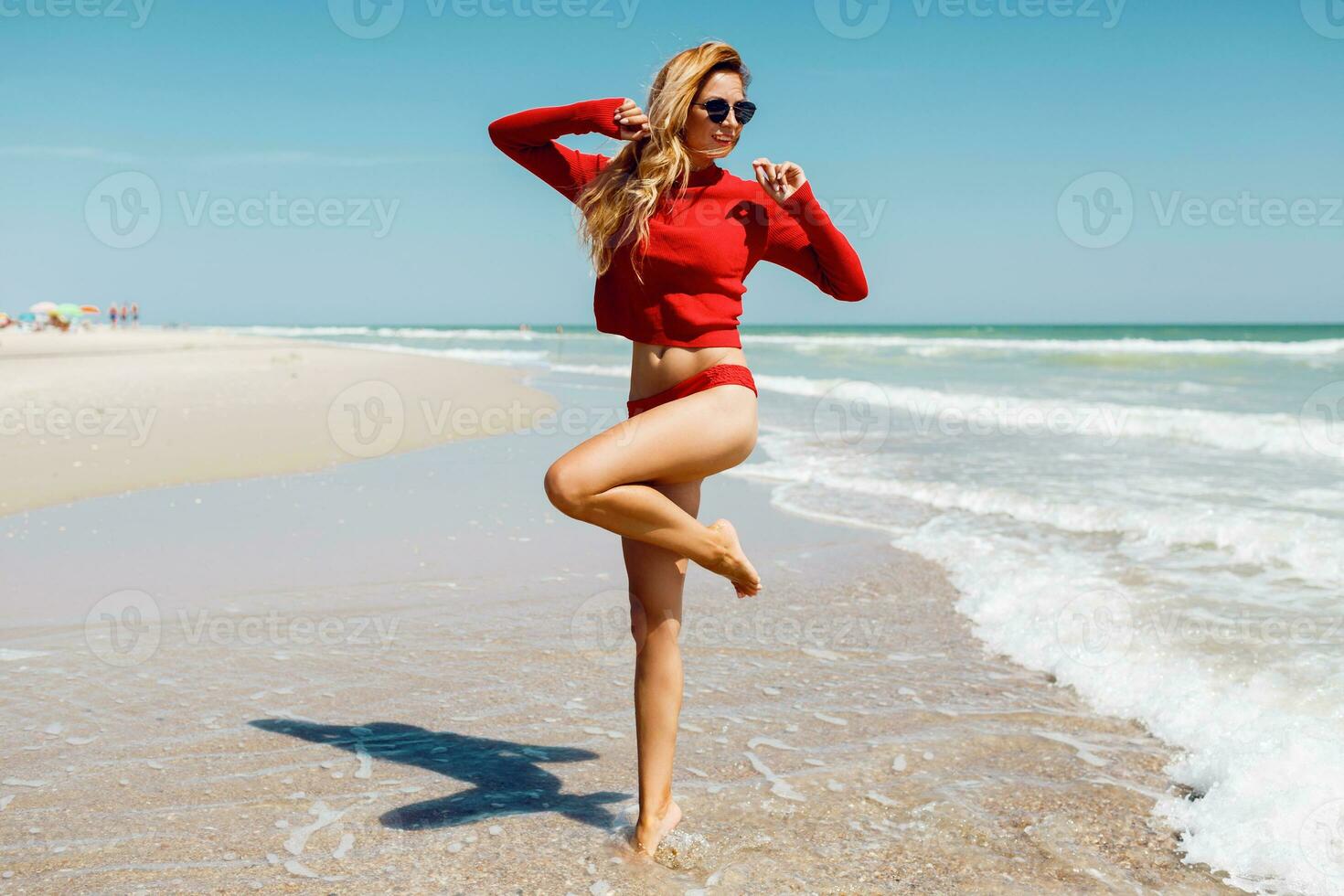Traveling blond woman jumping with happy expression , turns around in water. Ocean waves background. Wearing red bikini. Full length. Summer vacation. photo