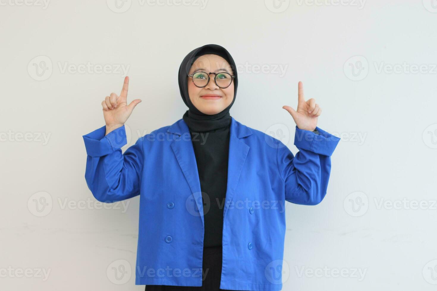 Beautiful young Asian Muslim woman in glasses, hijab and wearing blue blazer smiling confident and happy with both hands pointing up photo