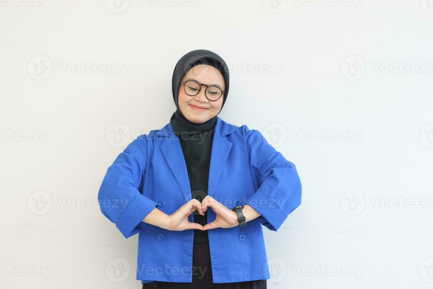 beautiful young asian woman wearing glasses, hijab and wearing blue blazer gesturing love sign with her hand photo