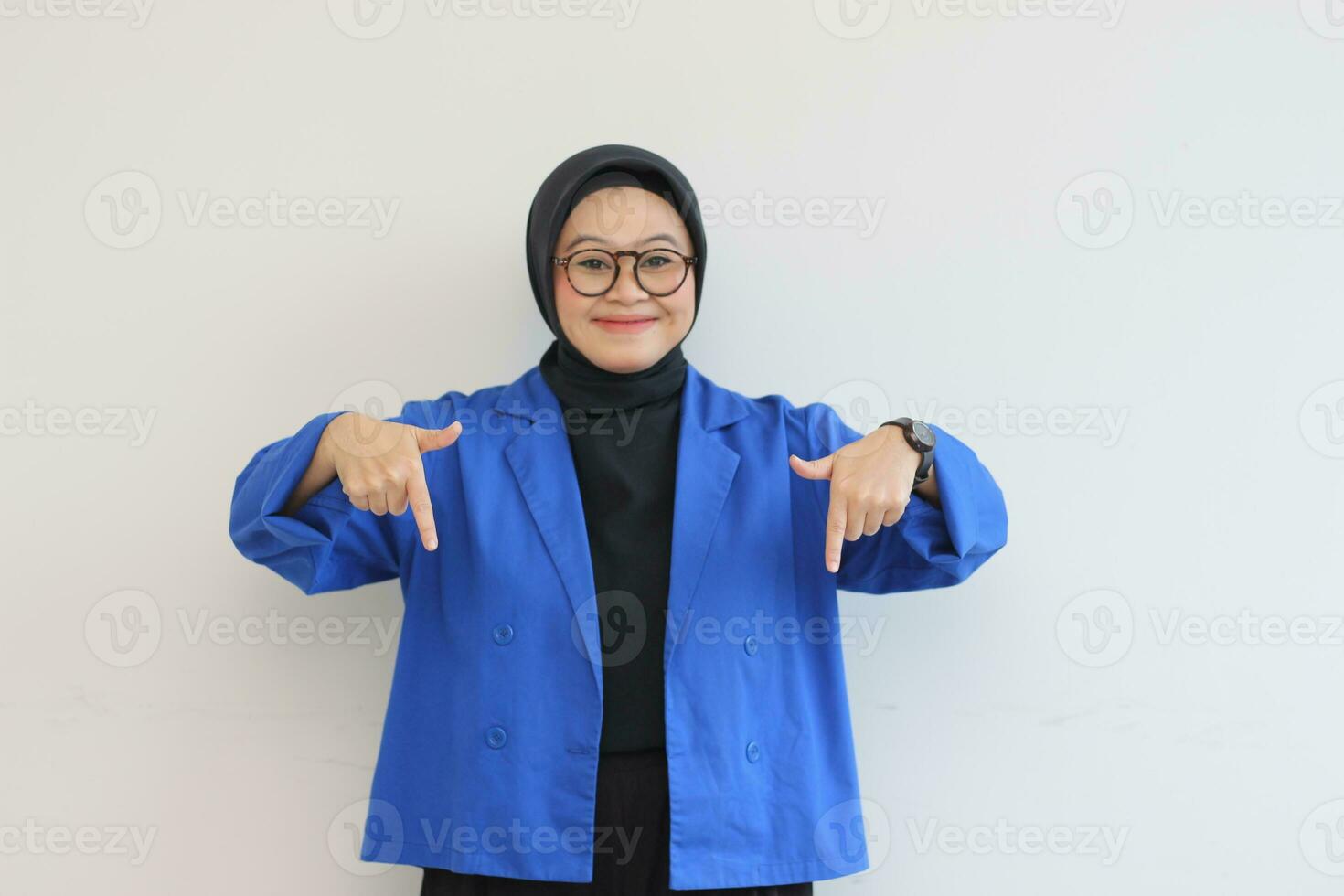 Beautiful young Asian Muslim woman in glasses, hijab and wearing blue blazer smiling confident and happy with both hands pointing down photo