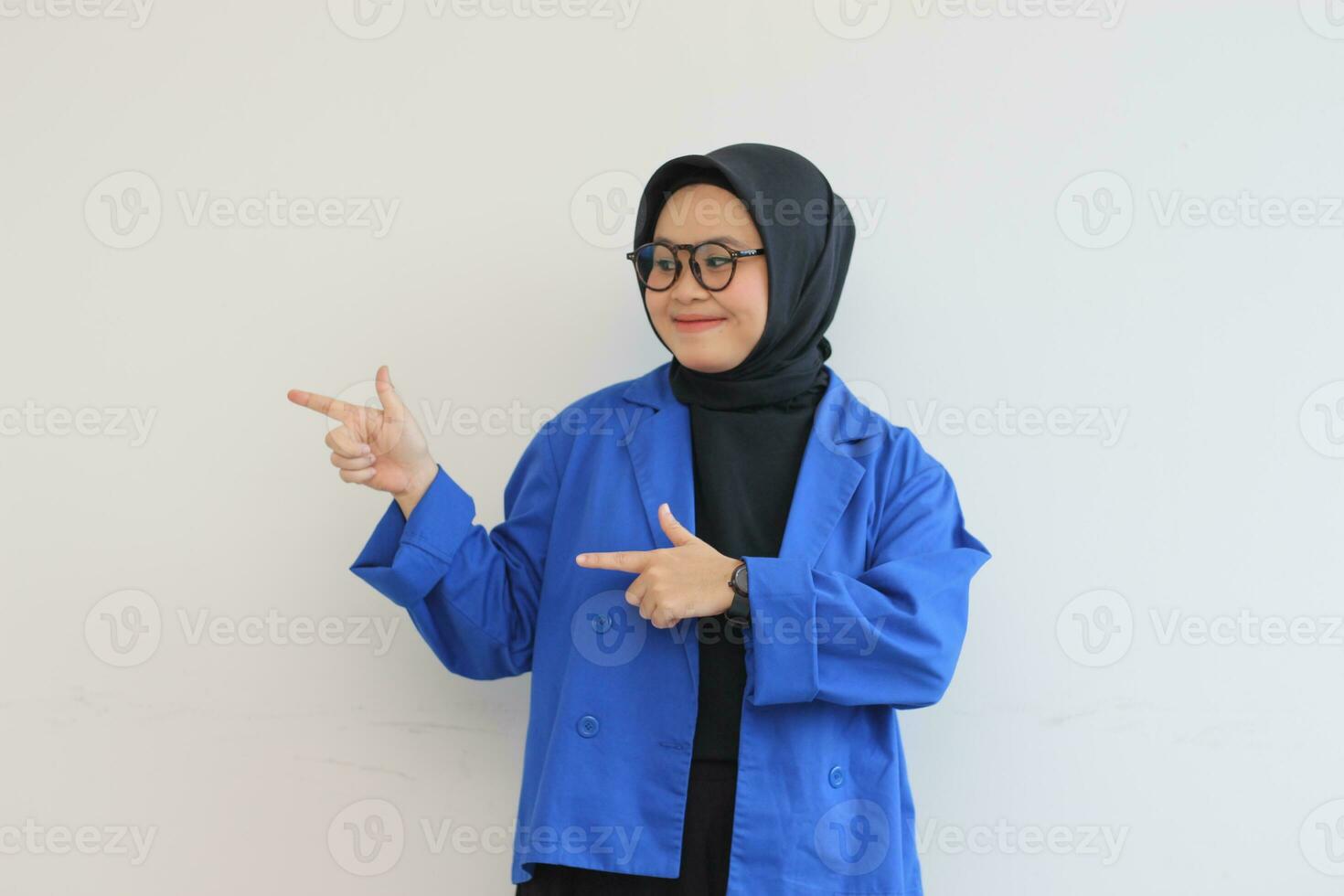 Beautiful young Asian Muslim woman in glasses, hijab and wearing blue blazer smiling confident and happy with both hands pointing sideways photo