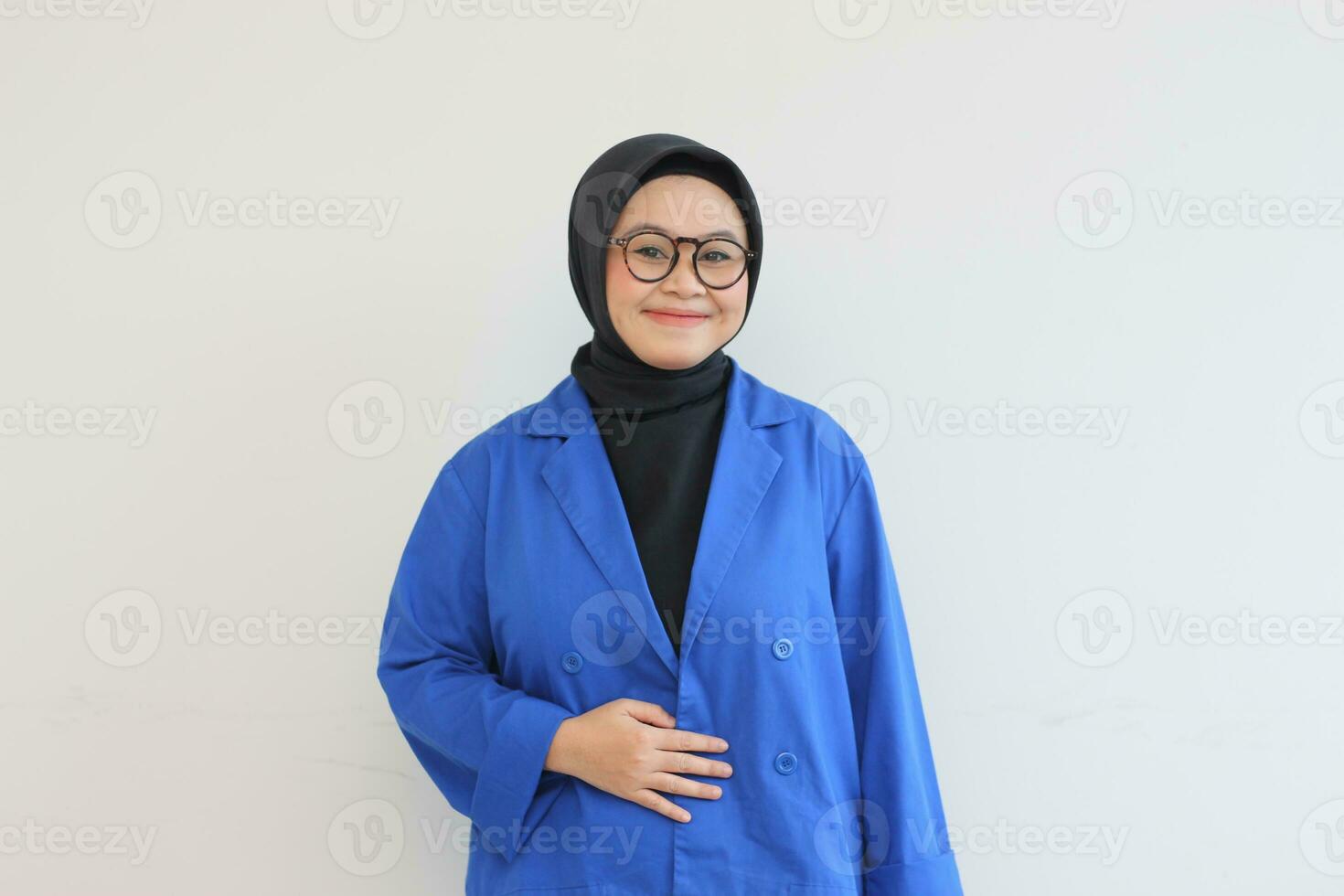 Beautiful young Asian Muslim woman in glasses, hijab and wearing blue blazer smiling confident and happy with hand on belly photo