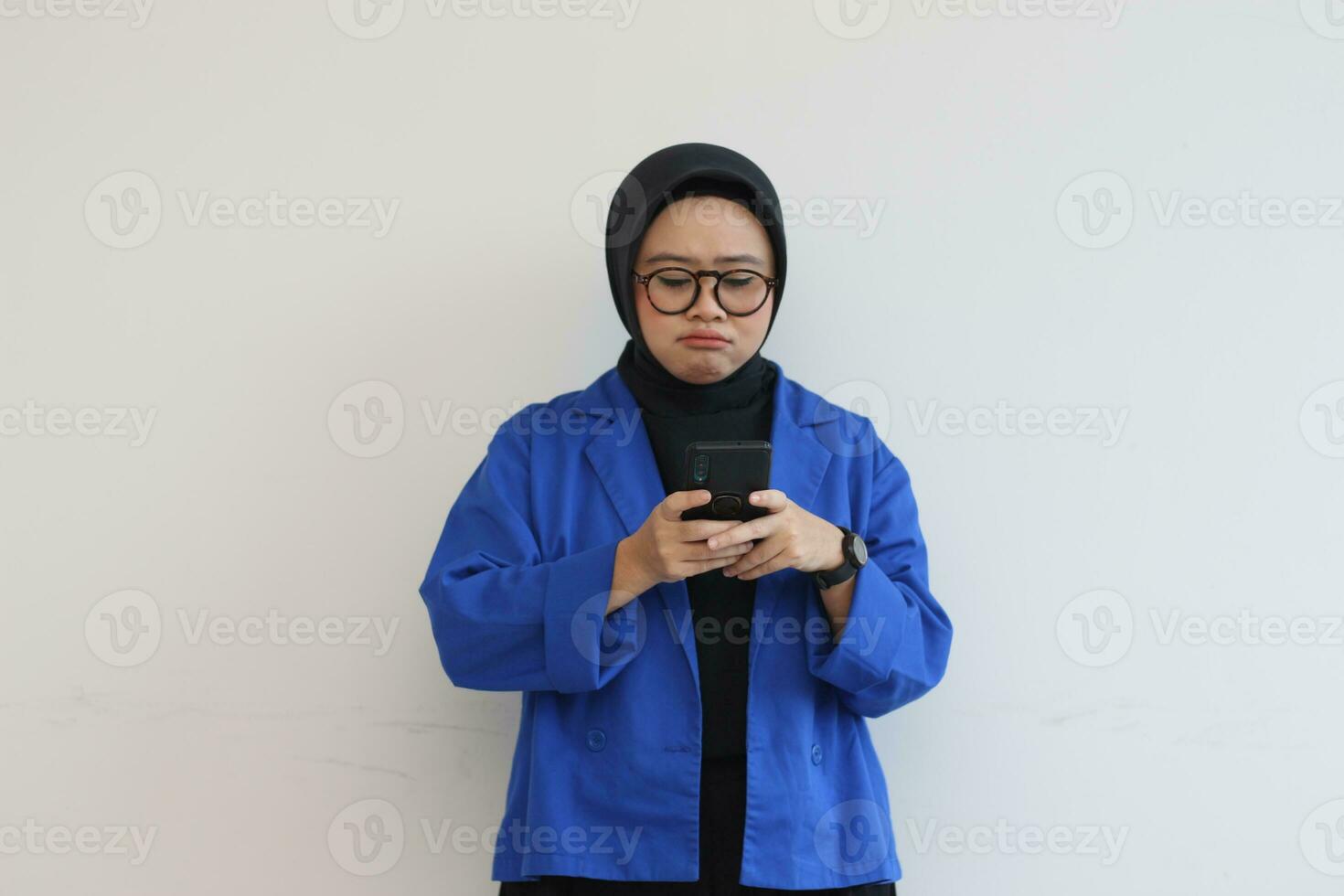 beautiful young asian woman in glasses, hijab and wearing blue blazer looking at her mobile phone with sullen face photo