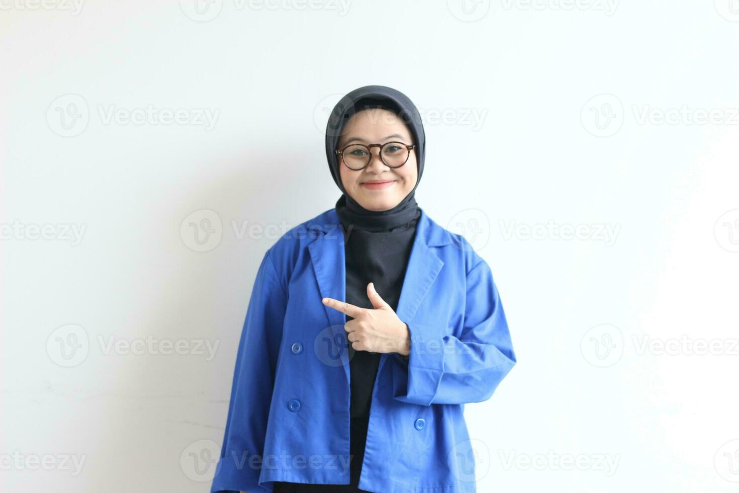 Beautiful young Asian Muslim woman in glasses, hijab and wearing blue blazer smiling confident and happy with both hands pointing sideways photo