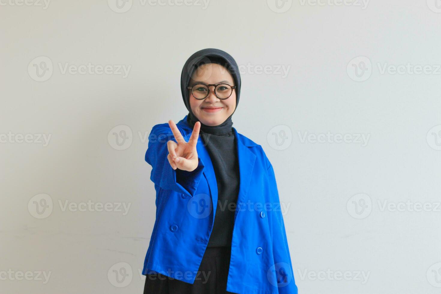 Beautiful young Asian Muslim woman, wearing glasses and blue blazer gesturing peace symbol photo