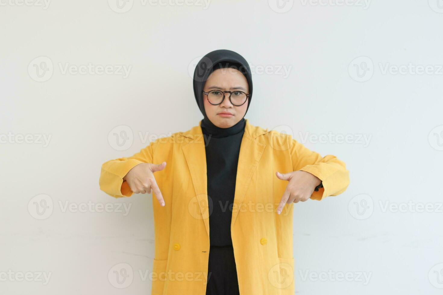 Beautiful young Asian Muslim woman in glasses, hijab and wearing yellow blazer sad expression with both hands pointing down photo