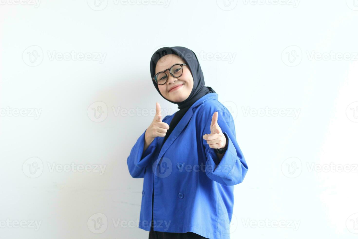 Beautiful young Asian Muslim woman in glasses, hijab and wearing blue blazer smiling confident and excited with both hands pointing forward photo