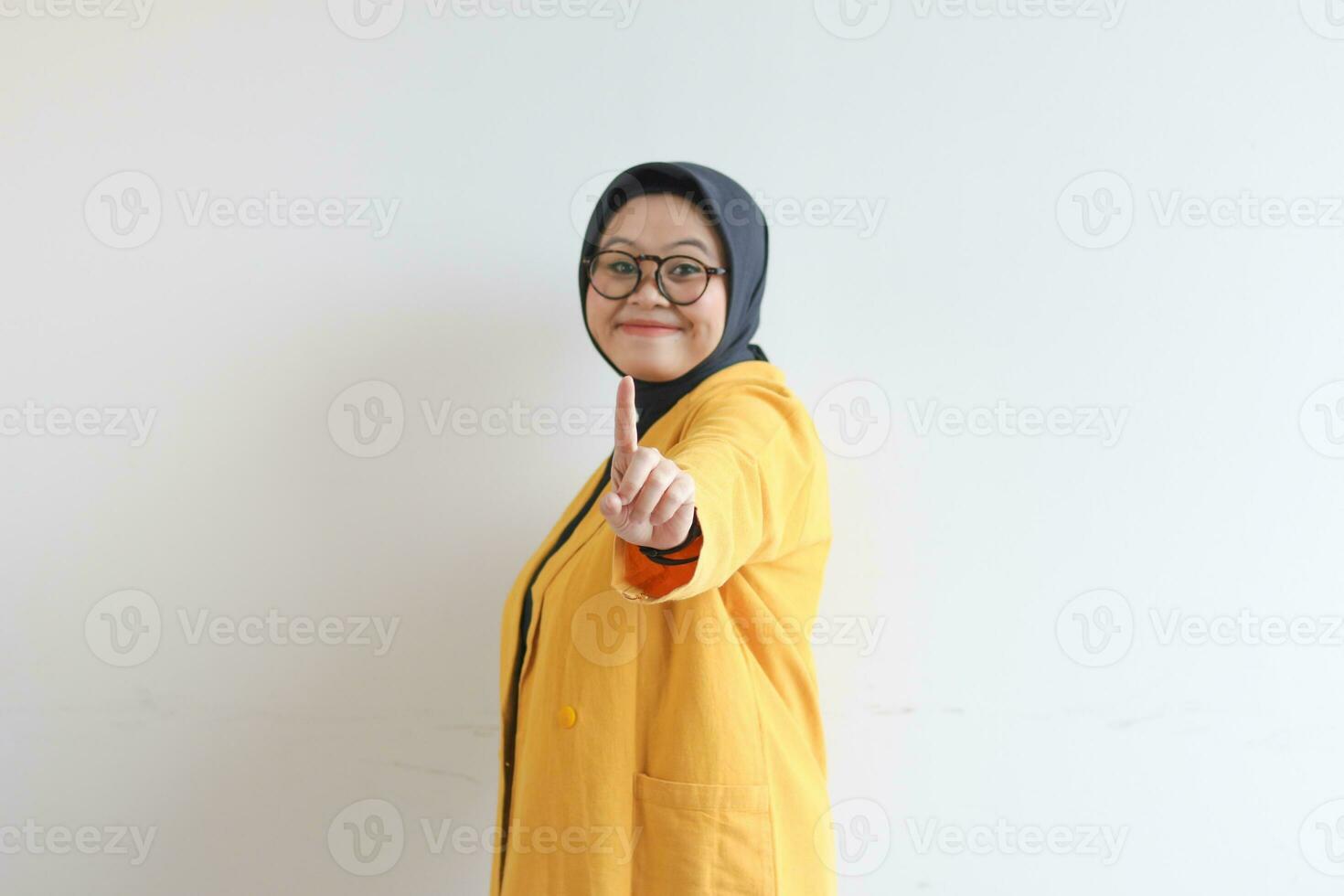 Young beautiful Asian Muslim woman, wearing glasses and yellow blazer gesturing number 1 sign hand with index finger photo