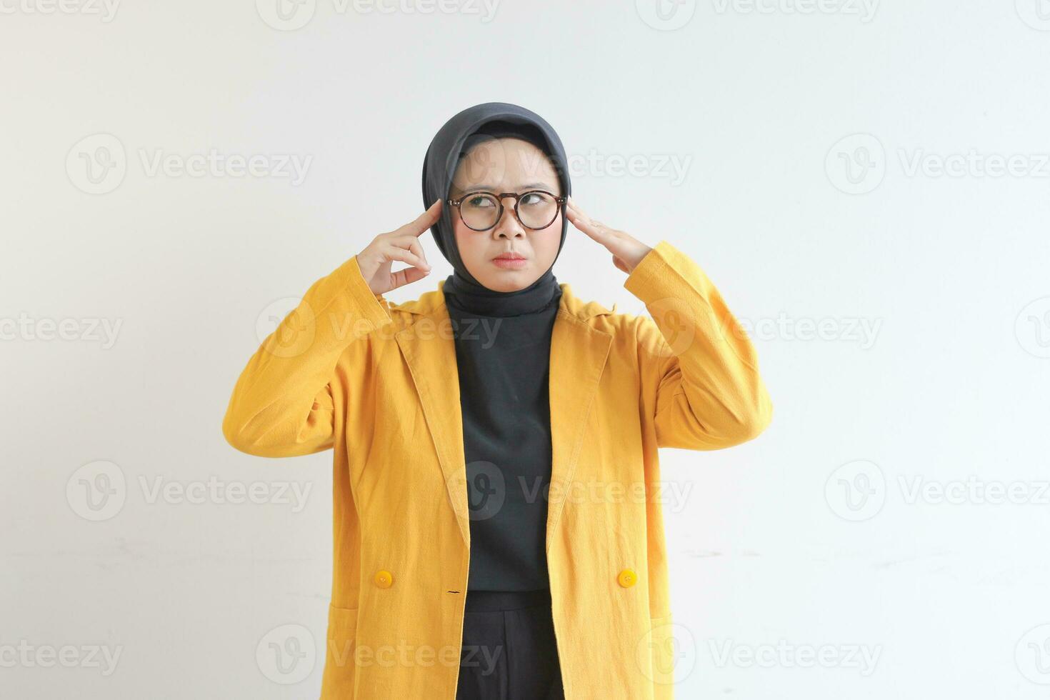 Beautiful young Asian Muslim woman, wearing glasses and yellow blazer is scratching head with confused expression photo