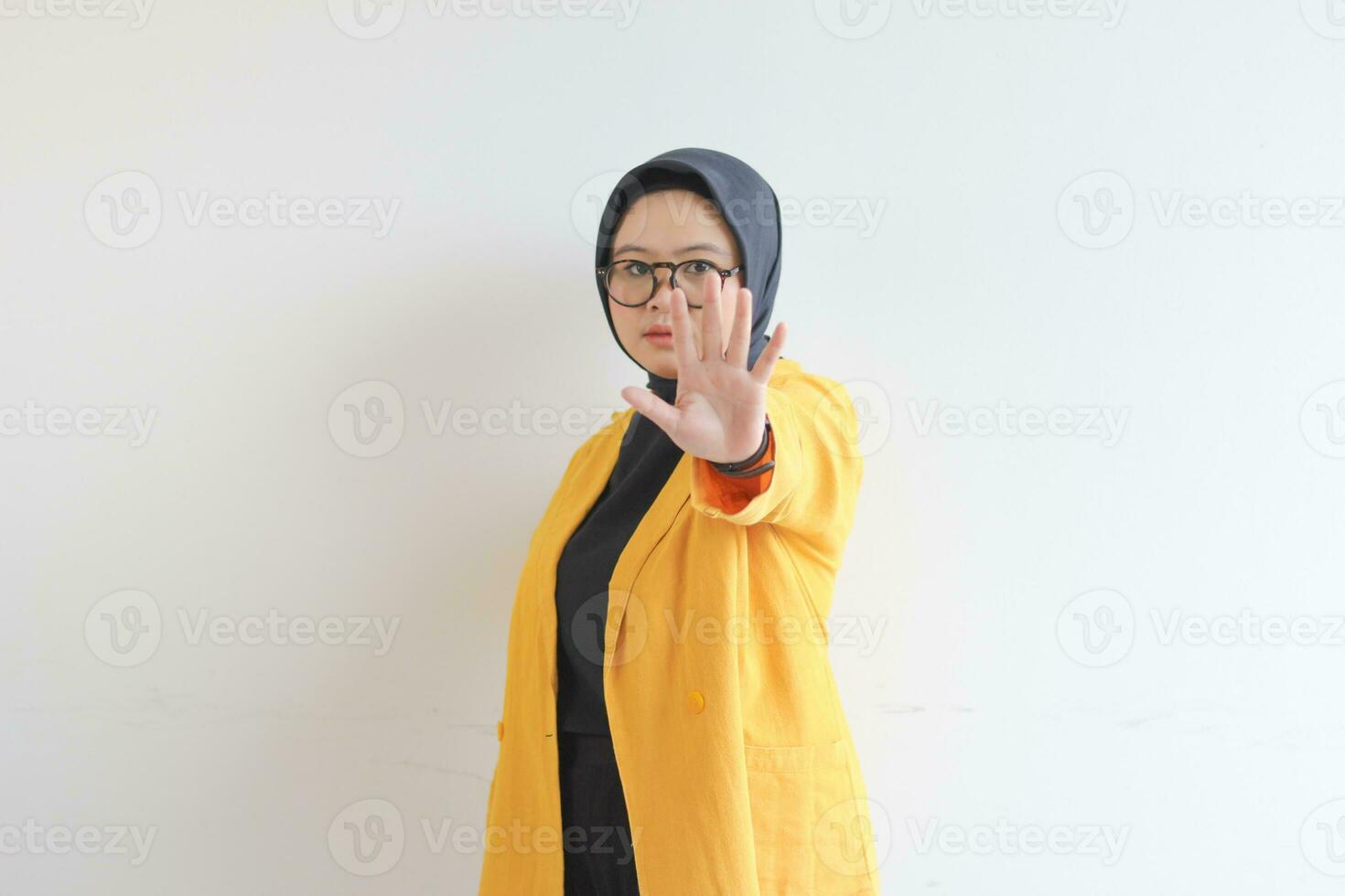 Beautiful young Asian Muslim woman, wearing glasses and yellow blazer showing stop gesture photo